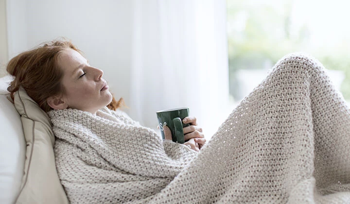 Frau mit Tee unter einer Decke Verschiedene Maßnahmen eignen sich, um eine Erkältung vorbeugen zu können
